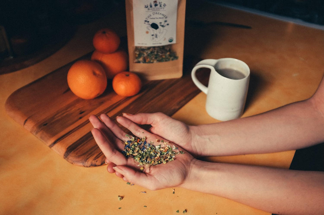 A person is using both hands to hold a scooping of loose leaf herbal tea in their palms. A cutting board is in the background with clementines and the bag of herbal tea on it. A white mug of freshly brewed tea is placed next to the cutting board.